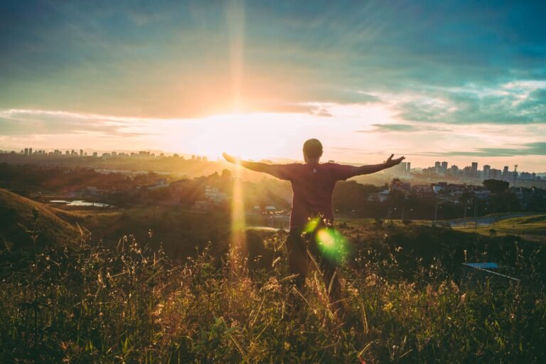 A person with outstretched arms on a hillside overlooking a city in warm sunlight.