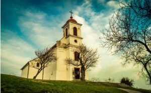 A white church building with a bell tower and cross beneath a cloudy blue sky.