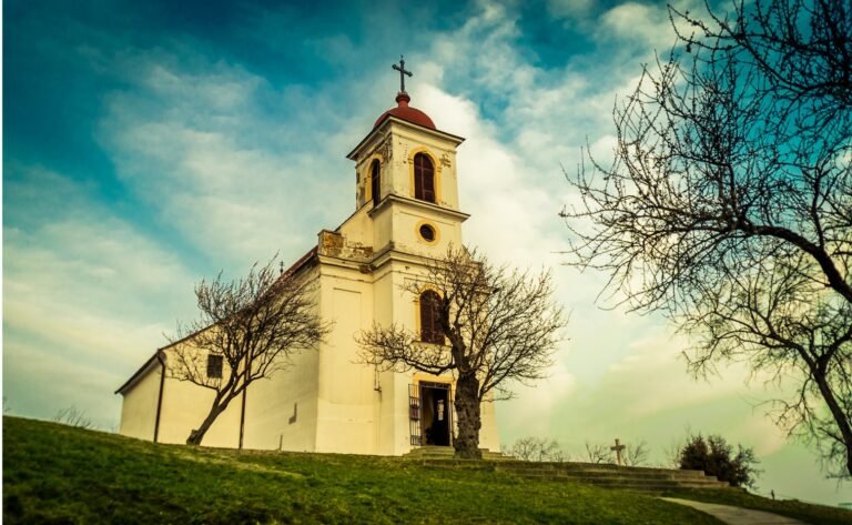A white church building with a bell tower and cross beneath a cloudy blue sky.