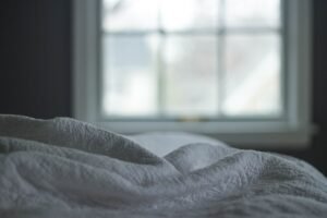 Rumpled gray bedcovers beside a softly lit window.