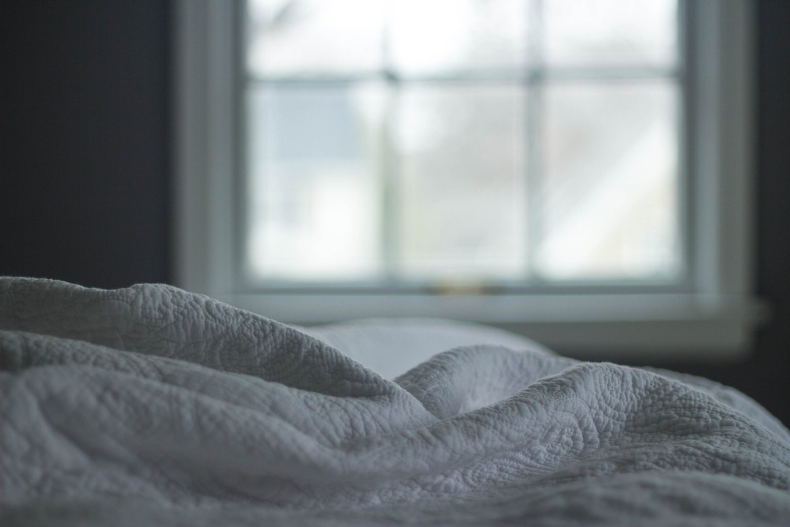 Rumpled gray bedcovers beside a softly lit window.